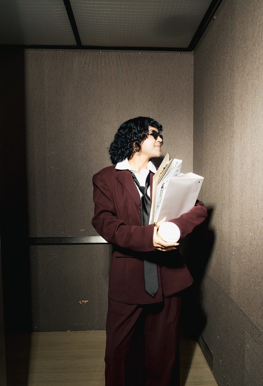 Annie in an elevator holding files and coffee