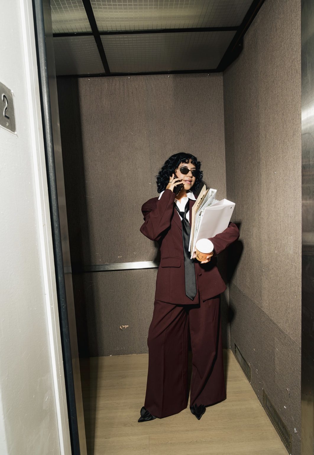 Annie on a phone call in an elevator with files and coffee
